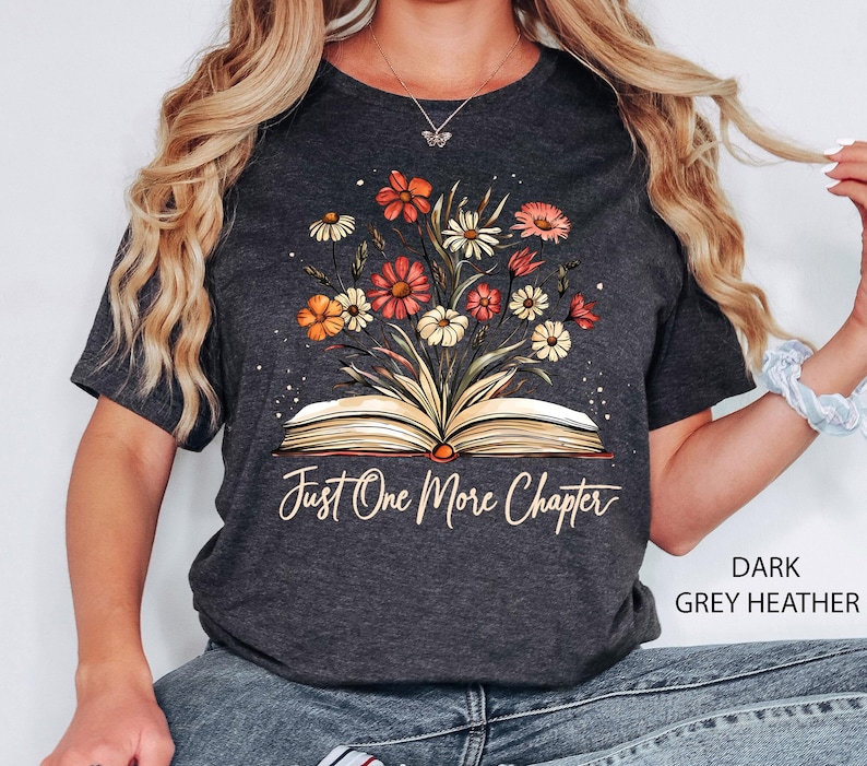 以下が含まれることがあります： ダークグレーヘザーのTシャツに、花柄のデザインと「Just One More Chapter」という文字が書かれた開いた本が描かれています。