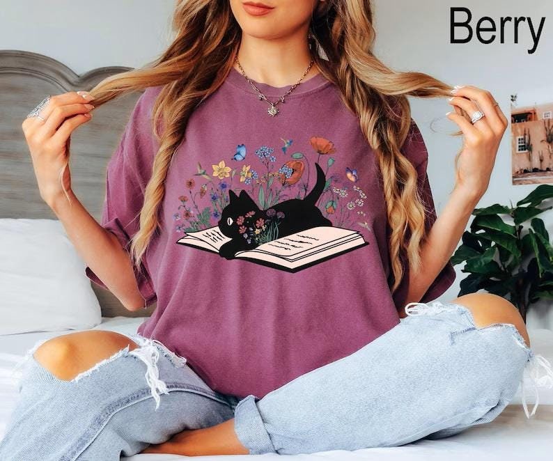 May include: A purple t-shirt with a black cat reading a book surrounded by flowers. The cat is sitting on an open book with the text "Berry" in the top right corner of the image.