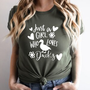 May include: Olive green t-shirt with the text "Just a GIRL WHO LOVES Ducks" in white. The design includes a duck silhouette, hearts, and flower graphics. The shirt is made of a soft material.