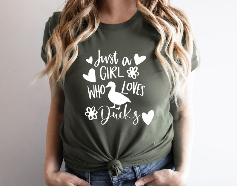 May include: Olive green t-shirt with the text "Just a GIRL WHO LOVES Ducks" in white. The design includes a duck silhouette, hearts, and flower graphics. The shirt is made of a soft material.