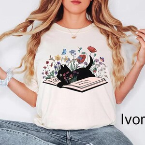 May include: A white t-shirt with a black cat reading a book surrounded by flowers. The cat is laying on the book with its paws on the pages. The flowers are in shades of pink, purple, blue, yellow, and green. The text "Ivory" is visible in the bottom right corner of the image.