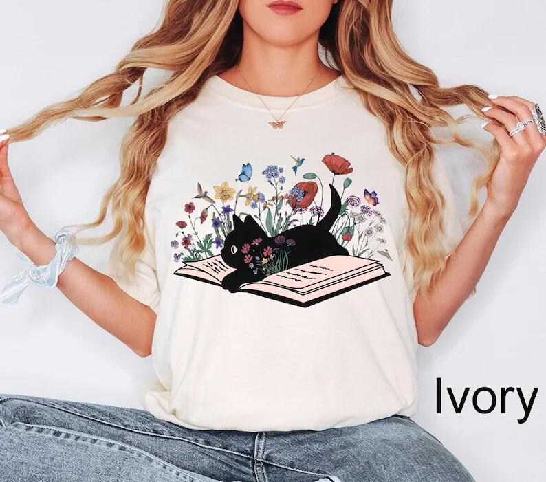 May include: A white t-shirt with a black cat reading a book surrounded by flowers. The cat is laying on the book with its paws on the pages. The flowers are in shades of pink, purple, blue, yellow, and green. The text "Ivory" is visible in the bottom right corner of the image.