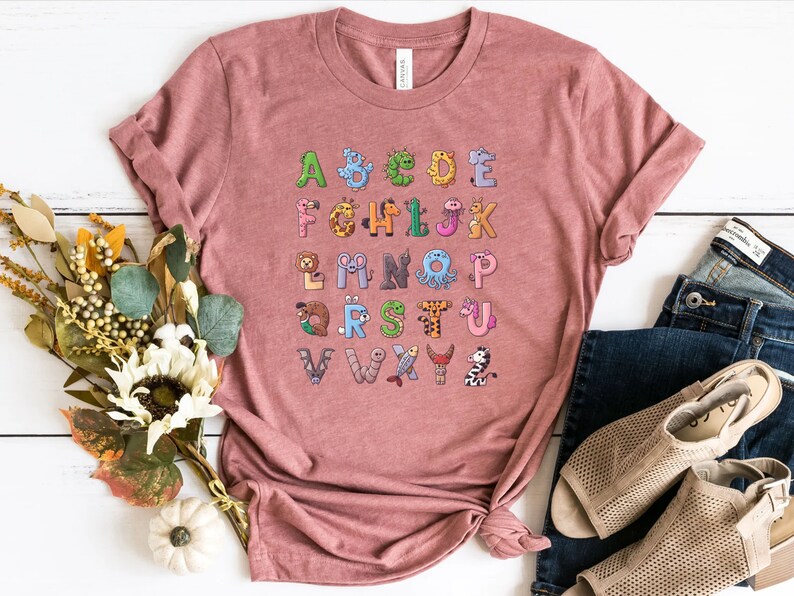 May include: A pink t-shirt with a colorful alphabet printed on the front. Each letter is decorated with a different animal illustration.
