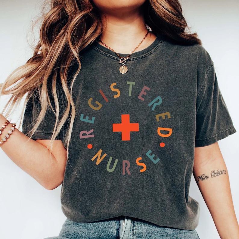 Puede incluir: Una camiseta gris oscuro con un gr&aacute;fico circular de colores que dice "REGISTERED NURSE" con una cruz roja en el centro.