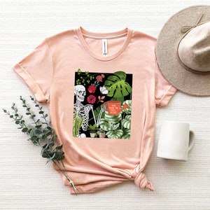 May include: A pink t-shirt with a graphic of a skeleton holding a watering can with the text "You make me feel alive". The graphic also includes plants and flowers.