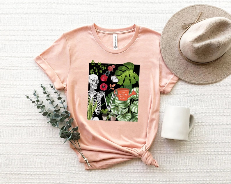 May include: A pink t-shirt with a graphic of a skeleton holding a watering can with the text "You make me feel alive". The graphic also includes plants and flowers.