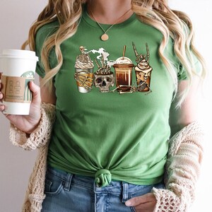 K&ouml;nnte beinhalten: Gr&uuml;nes T-Shirt mit einem Motiv von vier Kaffeetassen mit Skeletten und Sch&auml;deln. Die Tassen sind mit Schlagsahne und Schokoladensirup verziert. Der Text "Coffee & Bones" ist auf dem T-Shirt aufgedruckt.