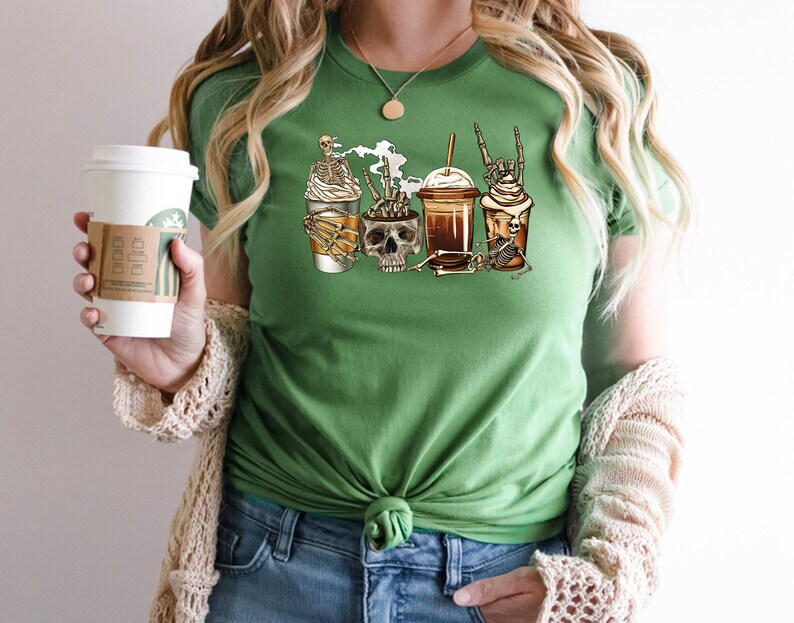 K&ouml;nnte beinhalten: Gr&uuml;nes T-Shirt mit einem Motiv von vier Kaffeetassen mit Skeletten und Sch&auml;deln. Die Tassen sind mit Schlagsahne und Schokoladensirup verziert. Der Text "Coffee & Bones" ist auf dem T-Shirt aufgedruckt.