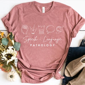 May include: A pink t-shirt with a white graphic design featuring icons related to speech and language pathology. The text "Speech - Language PATHOLOGY" is printed below the icons.