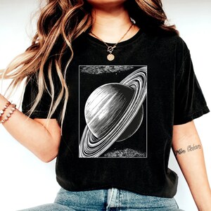 May include: A black t-shirt with a white graphic of Saturn, a planet with rings, on the front. The graphic is in a vintage style.