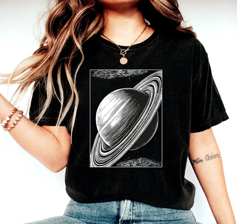 May include: A black t-shirt with a white graphic of Saturn, a planet with rings, on the front. The graphic is in a vintage style.