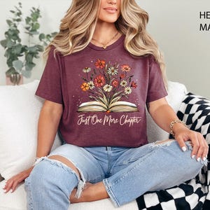 以下が含まれることがあります： ヘザーマロンのTシャツに、開いた本から花が咲いているグラフィックがプリントされています。本の下に「Just One More Chapter」というテキストがプリントされています。