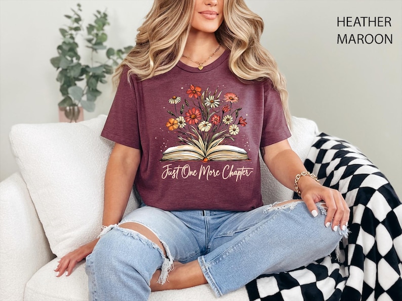 以下が含まれることがあります： ヘザーマロンのTシャツに、開いた本から花が咲いているグラフィックがプリントされています。本の下に「Just One More Chapter」というテキストがプリントされています。
