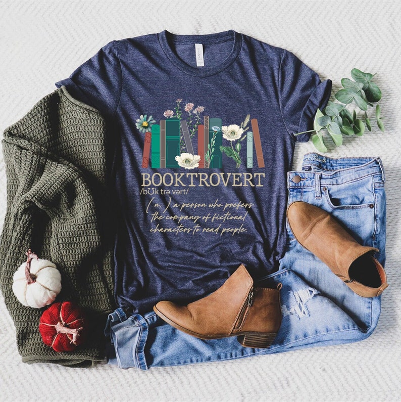 Puede incluir: Una camiseta azul marino con un gr&aacute;fico de libros apilados con flores y el texto "BOOKTROVERT /bUk trə vərt/ (n) a person who prefers the company of fictional characters to read people."