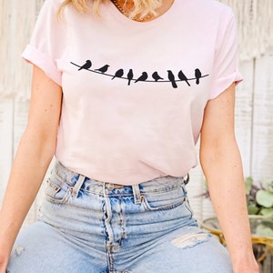 May include: A pink t-shirt with a graphic of a line of black birds on a wire. The shirt has short sleeves and a crew neck.