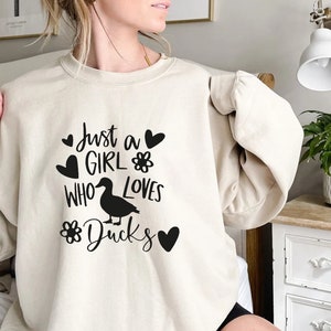 May include: Beige sweatshirt with the text "Just a GIRL WHO LOVES Ducks" in black. The design includes a duck silhouette, hearts, and flower graphics. The sweatshirt is a casual, comfortable style.