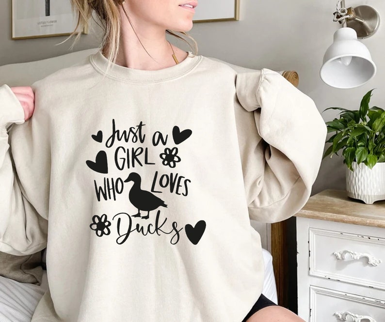 May include: Beige sweatshirt with the text "Just a GIRL WHO LOVES Ducks" in black. The design includes a duck silhouette, hearts, and flower graphics. The sweatshirt is a casual, comfortable style.