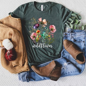 May include: A dark green t-shirt featuring a watercolour floral design and the word "wildflower" printed on the front. The shirt is laid out on a white surface with a brown sweater, blue jeans, and brown boots.