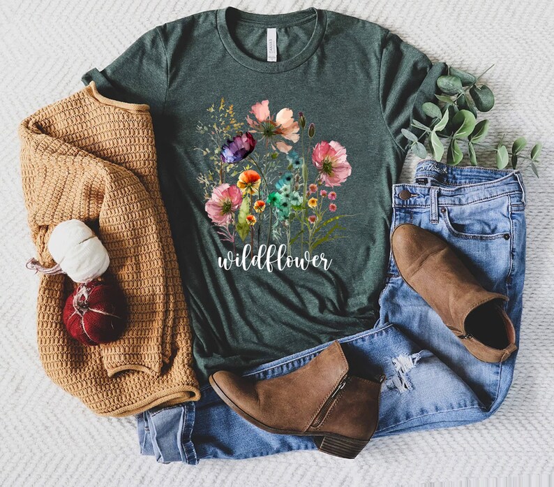 May include: A dark green t-shirt featuring a watercolour floral design and the word "wildflower" printed on the front. The shirt is laid out on a white surface with a brown sweater, blue jeans, and brown boots.