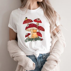May include: White t-shirt with a graphic of a cat sitting under red and white mushrooms with a butterfly.