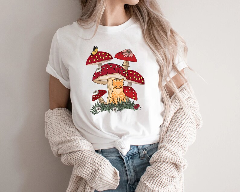 May include: White t-shirt with a graphic of a cat sitting under red and white mushrooms with a butterfly.