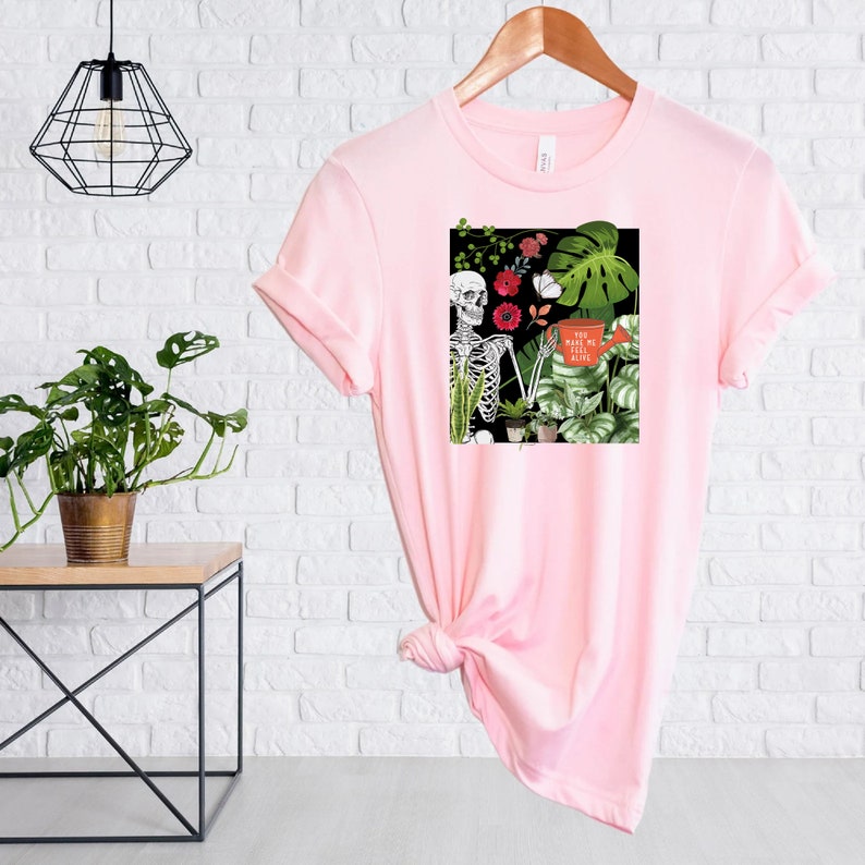 May include: Pink t-shirt with a graphic of a skeleton holding a watering can and surrounded by plants. The text on the graphic says "You make me feel alive".
