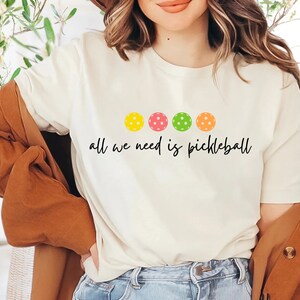 May include: White t-shirt with a graphic of four colorful pickleball paddles and the text "all we need is pickleball".