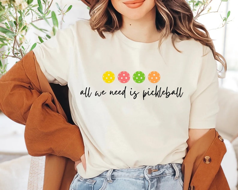 May include: White t-shirt with a graphic of four colorful pickleball paddles and the text "all we need is pickleball".