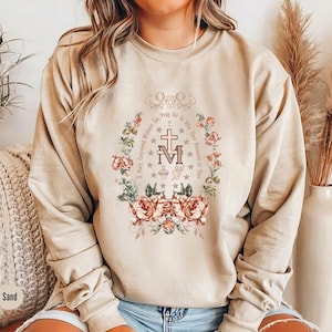 Op de afbeelding: Een beige sweatshirt met een bloemenontwerp en de tekst "Maria, ontvangen zonder zonde, bid voor ons die onze toevlucht tot u nemen." Het ontwerp bevat een kruis en de letters "M" en "M" die in elkaar zijn verweven.