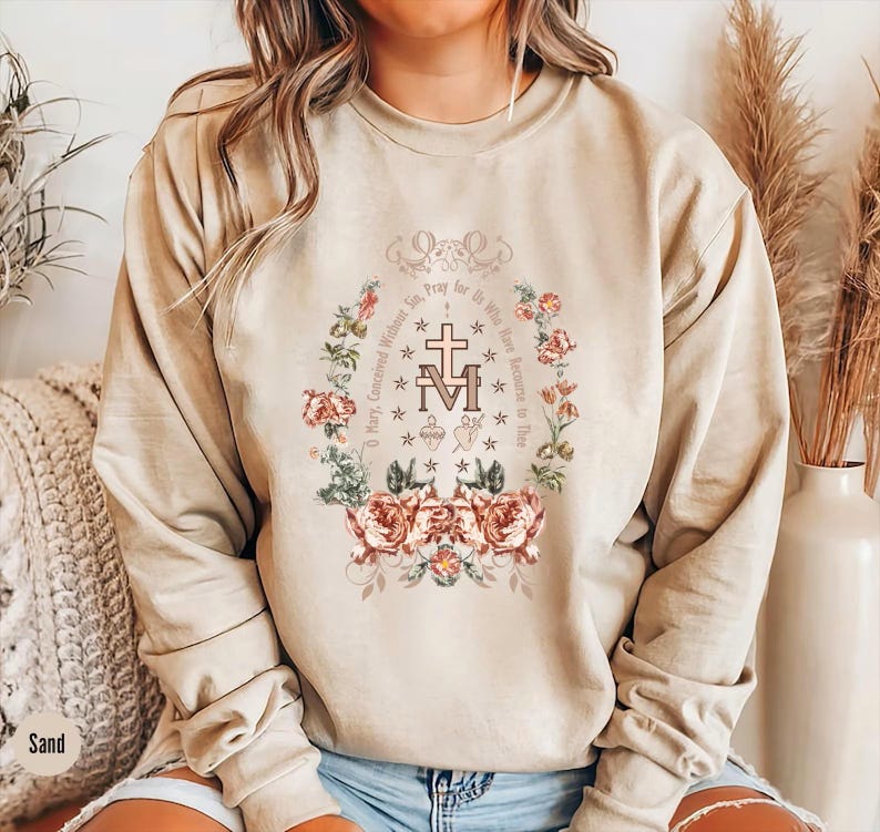 Op de afbeelding: Een beige sweatshirt met een bloemenontwerp en de tekst "Maria, ontvangen zonder zonde, bid voor ons die onze toevlucht tot u nemen." Het ontwerp bevat een kruis en de letters "M" en "M" die in elkaar zijn verweven.
