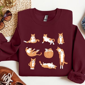K&ouml;nnte beinhalten: Ein maroonfarbener Pullover mit einem grafischen Druck von neun orangefarbenen und wei&szlig;en Katzen in verschiedenen Yoga-Posen.