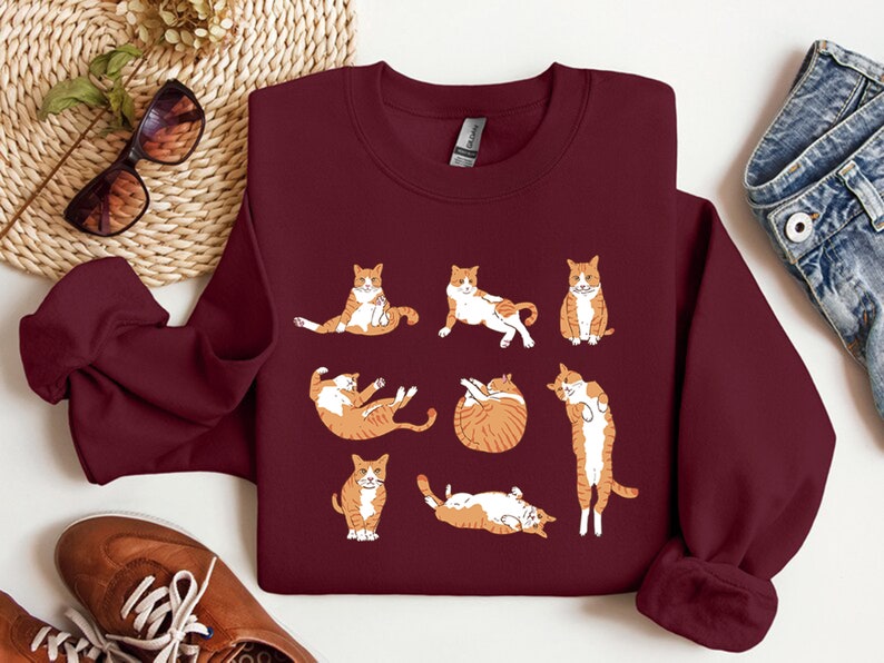 K&ouml;nnte beinhalten: Ein maroonfarbener Pullover mit einem grafischen Druck von neun orangefarbenen und wei&szlig;en Katzen in verschiedenen Yoga-Posen.