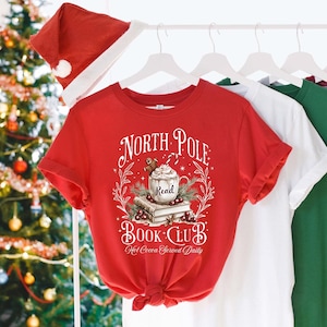 K&ouml;nnte beinhalten: Rotes T-Shirt mit einem wei&szlig;en Grafikdesign, auf dem "North Pole Book Club" steht, darunter eine Tasse hei&szlig;e Schokolade und B&uuml;cher. Der Text lautet au&szlig;erdem "Hot Cocoa Served Daily".