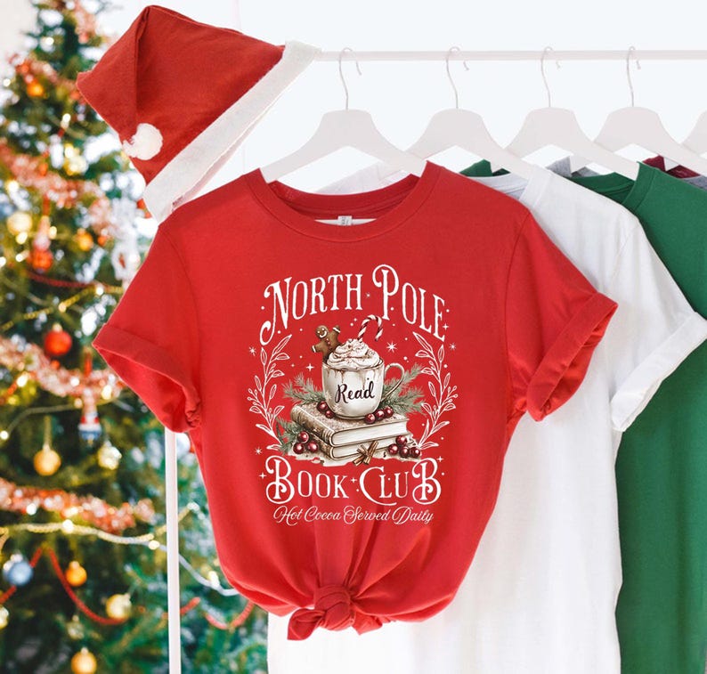 K&ouml;nnte beinhalten: Rotes T-Shirt mit einem wei&szlig;en Grafikdesign, auf dem "North Pole Book Club" steht, darunter eine Tasse hei&szlig;e Schokolade und B&uuml;cher. Der Text lautet au&szlig;erdem "Hot Cocoa Served Daily".