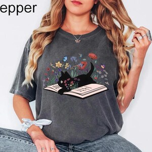 May include: A gray t-shirt with a black cat reading a book surrounded by flowers. The cat is looking at the book. The shirt has the word "Pepper" printed on the top left corner.