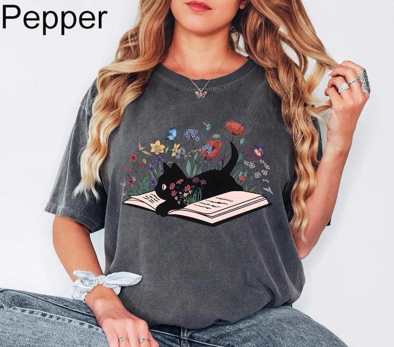 May include: A gray t-shirt with a black cat reading a book surrounded by flowers. The cat is looking at the book. The shirt has the word "Pepper" printed on the top left corner.