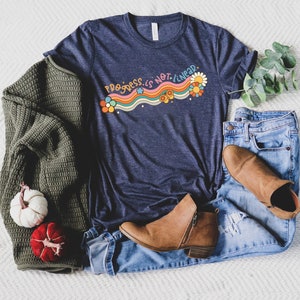 K&ouml;nnte beinhalten: Ein marineblaues T-Shirt mit einem Regenbogen-Design und dem Text "Progress is not linear" in Wei&szlig;. Das Shirt wird mit einem gr&uuml;nen Pullover, blauen Jeans und braunen Stiefeletten kombiniert.
