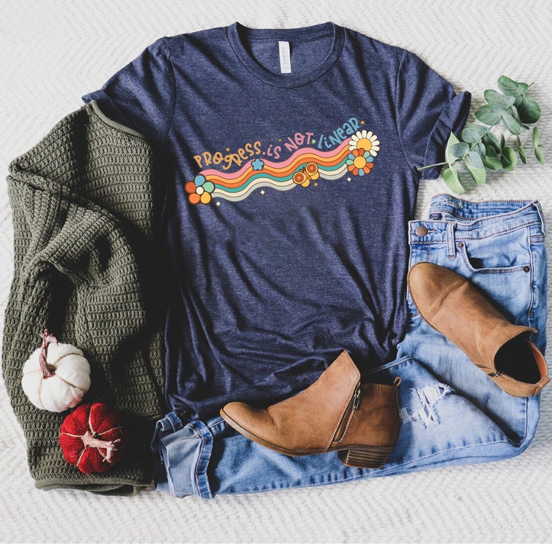 K&ouml;nnte beinhalten: Ein marineblaues T-Shirt mit einem Regenbogen-Design und dem Text "Progress is not linear" in Wei&szlig;. Das Shirt wird mit einem gr&uuml;nen Pullover, blauen Jeans und braunen Stiefeletten kombiniert.