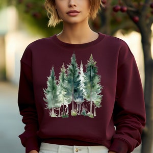 Puede incluir: Una sudadera color burdeos con un estampado de acuarela de una escena de bosque. Los &aacute;rboles son verdes y azules, y el fondo es verde claro.
