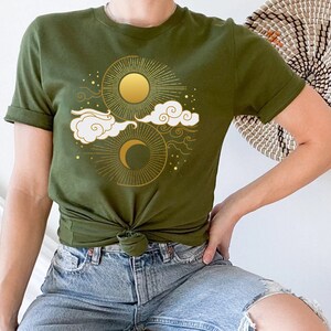 May include: A green t-shirt with a gold sun and moon design. The design features stylized clouds and stars.