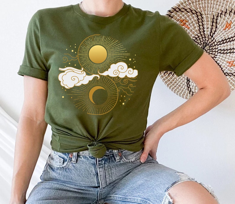 May include: A green t-shirt with a gold sun and moon design. The design features stylized clouds and stars.