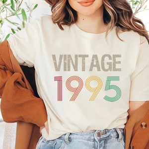 May include: A white t-shirt with the text "Vintage 1995" printed in a retro style with multi-colored stripes. The year is in a larger font than the word "Vintage".