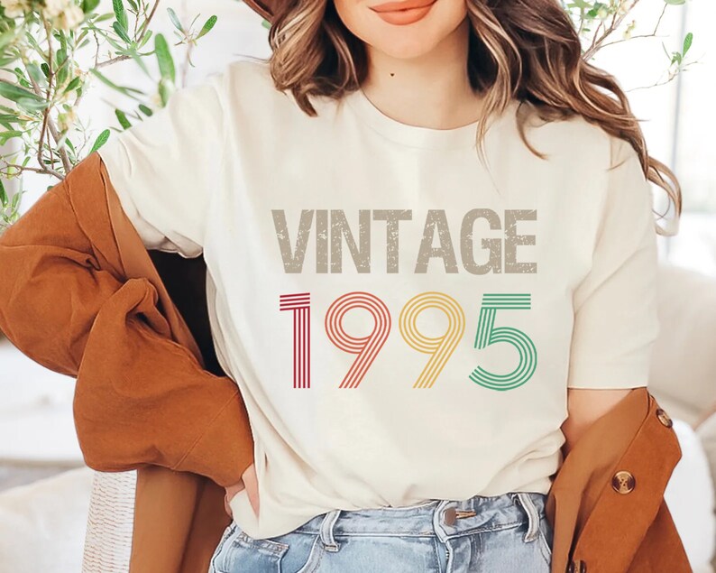 May include: A white t-shirt with the text "Vintage 1995" printed in a retro style with multi-colored stripes. The year is in a larger font than the word "Vintage".
