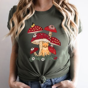 May include: A green t-shirt with a graphic of a cat sitting under a cluster of red and white mushrooms with white dots. The mushrooms have green stems and white flowers.