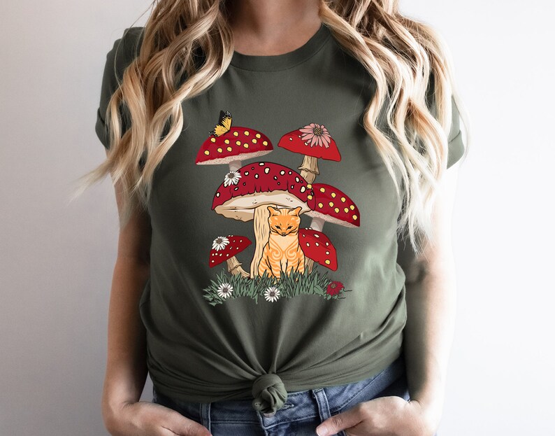 May include: A green t-shirt with a graphic of a cat sitting under a cluster of red and white mushrooms with white dots. The mushrooms have green stems and white flowers.