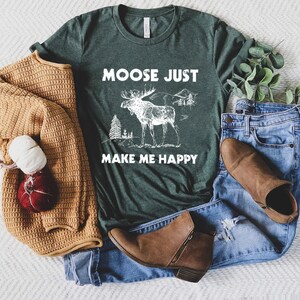 May include: Dark green t-shirt with the text "MOOSE JUST MAKE ME HAPPY" and a white graphic of a moose, trees, and mountains. The shirt is paired with blue jeans, brown boots, and a tan sweater.