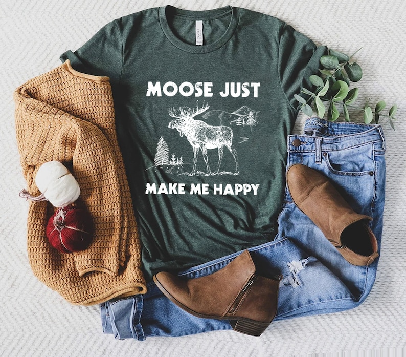 May include: Dark green t-shirt with the text "MOOSE JUST MAKE ME HAPPY" and a white graphic of a moose, trees, and mountains. The shirt is paired with blue jeans, brown boots, and a tan sweater.