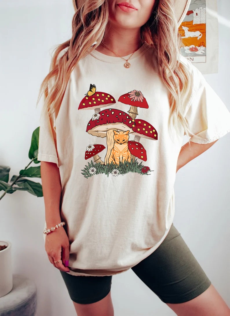 May include: A cream-colored t-shirt with a graphic of a cat sitting under a cluster of red and white mushrooms. The mushrooms have white spots and are surrounded by green grass and white flowers.
