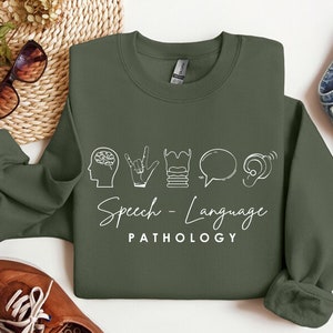 May include: A green sweatshirt with white text that reads "Speech - Language PATHOLOGY". The text is accompanied by five icons representing speech therapy: a brain, a hand making the sign for "I love you", a stack of books, a speech bubble, and an ear.