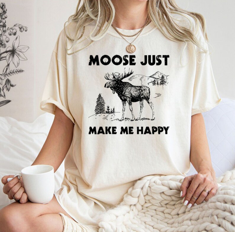 May include: Cream-colored t-shirt with the text "MOOSE JUST MAKE ME HAPPY" in black. The graphic features a detailed black and white illustration of a moose with antlers, trees, and mountains.